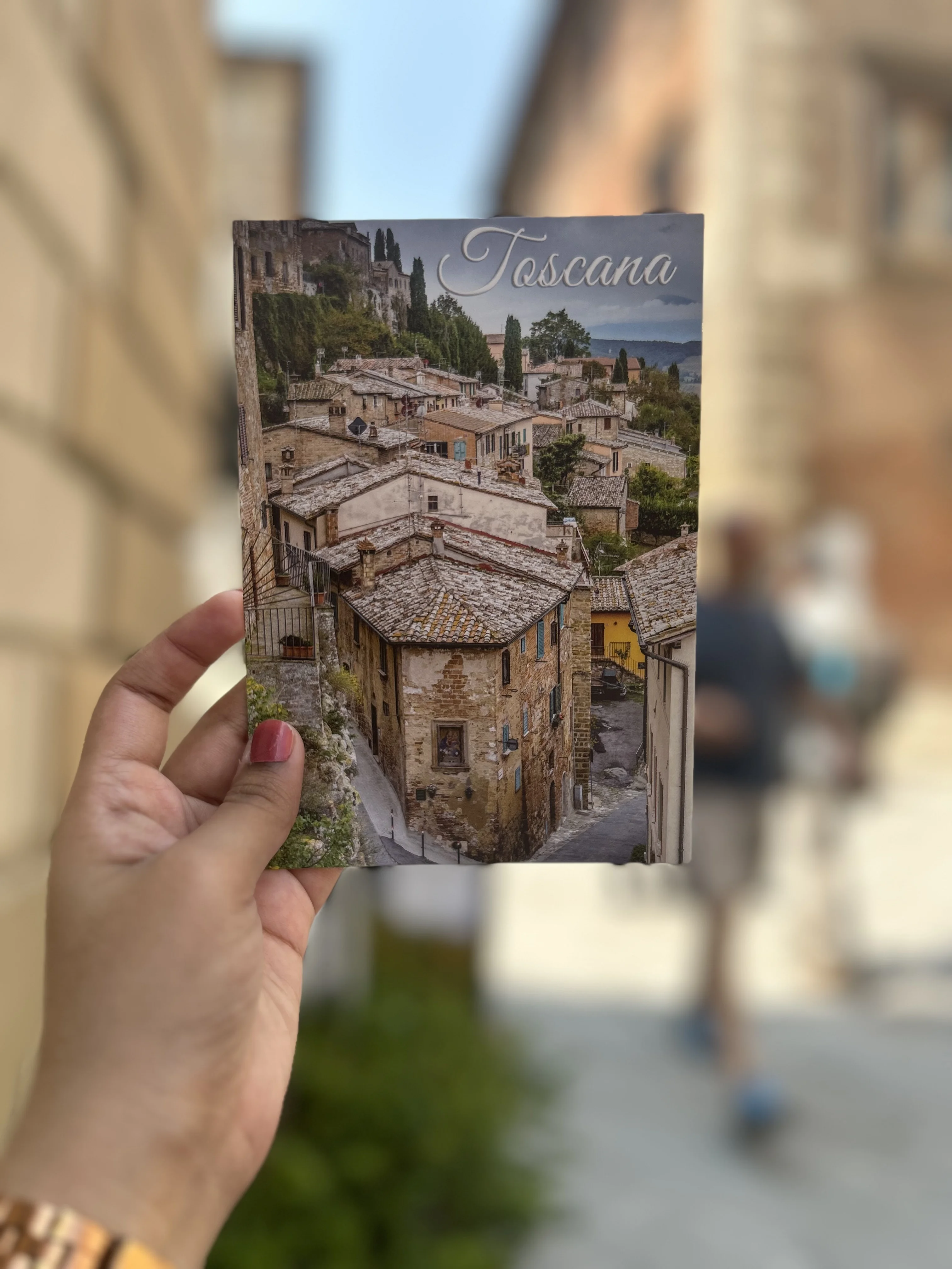 Hand holding a Toscana postcard against the blurred streets of Montepulciano